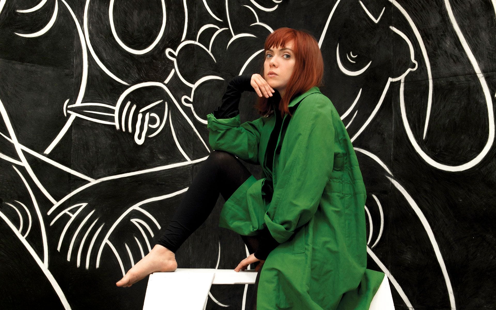Person in green coat seated in front of a black and white abstract mural.