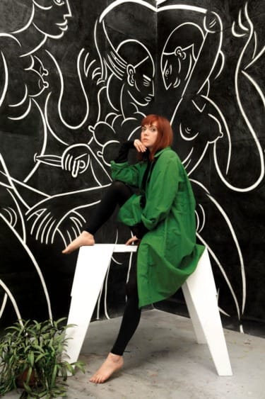 Woman in a green coat sitting on a chair in front of a mural with white abstract figures, plant on the floor nearby.