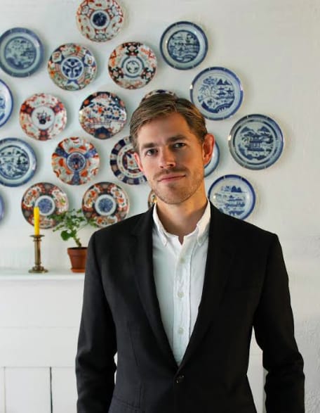 Person in a suit stands in front of decorative plates on a wall.