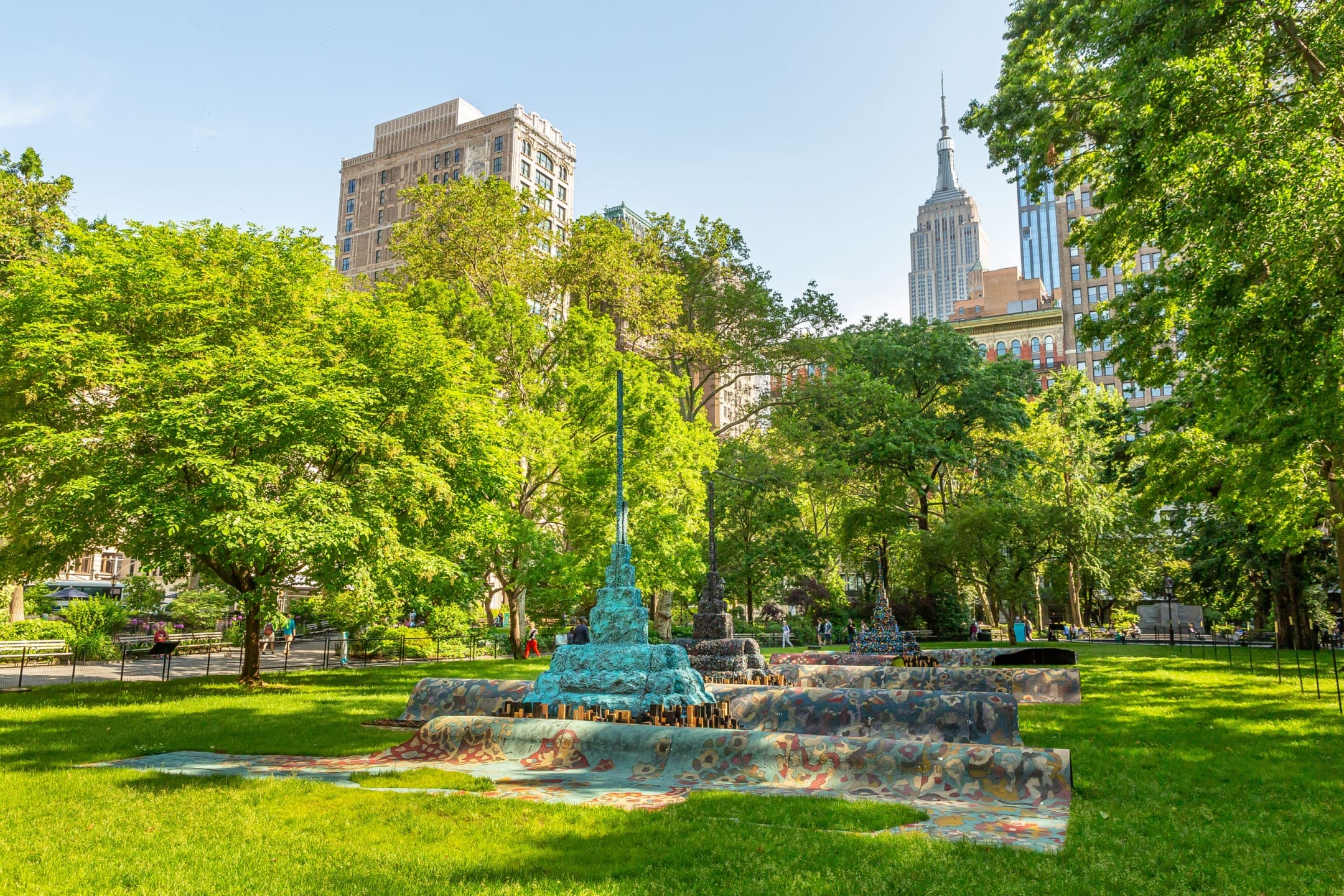 Colorful outdoor art installation in a park surrounded by lush greenery and tall buildings in the background.