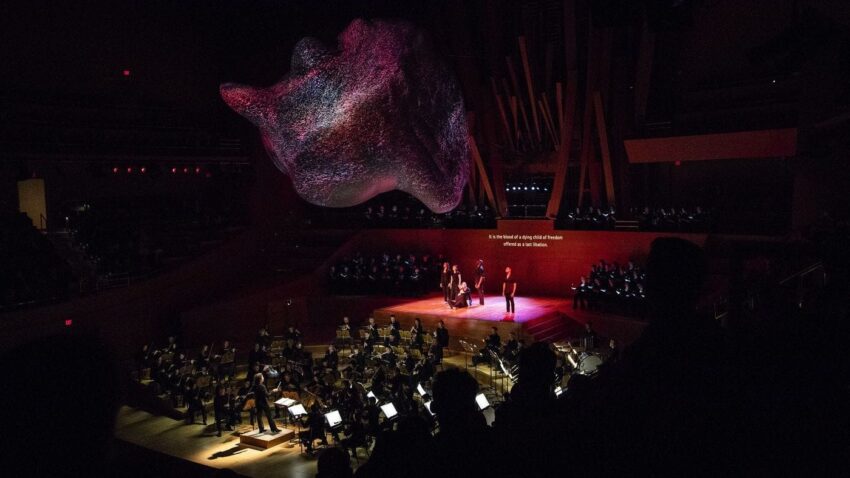 Orchestra performing on stage with abstract, illuminated art installation overhead in a dimly lit concert hall.