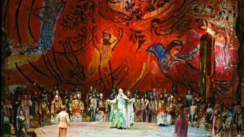 Colorful stage performance with a large cast in vibrant costumes and a vivid, artistic backdrop depicting various figures.