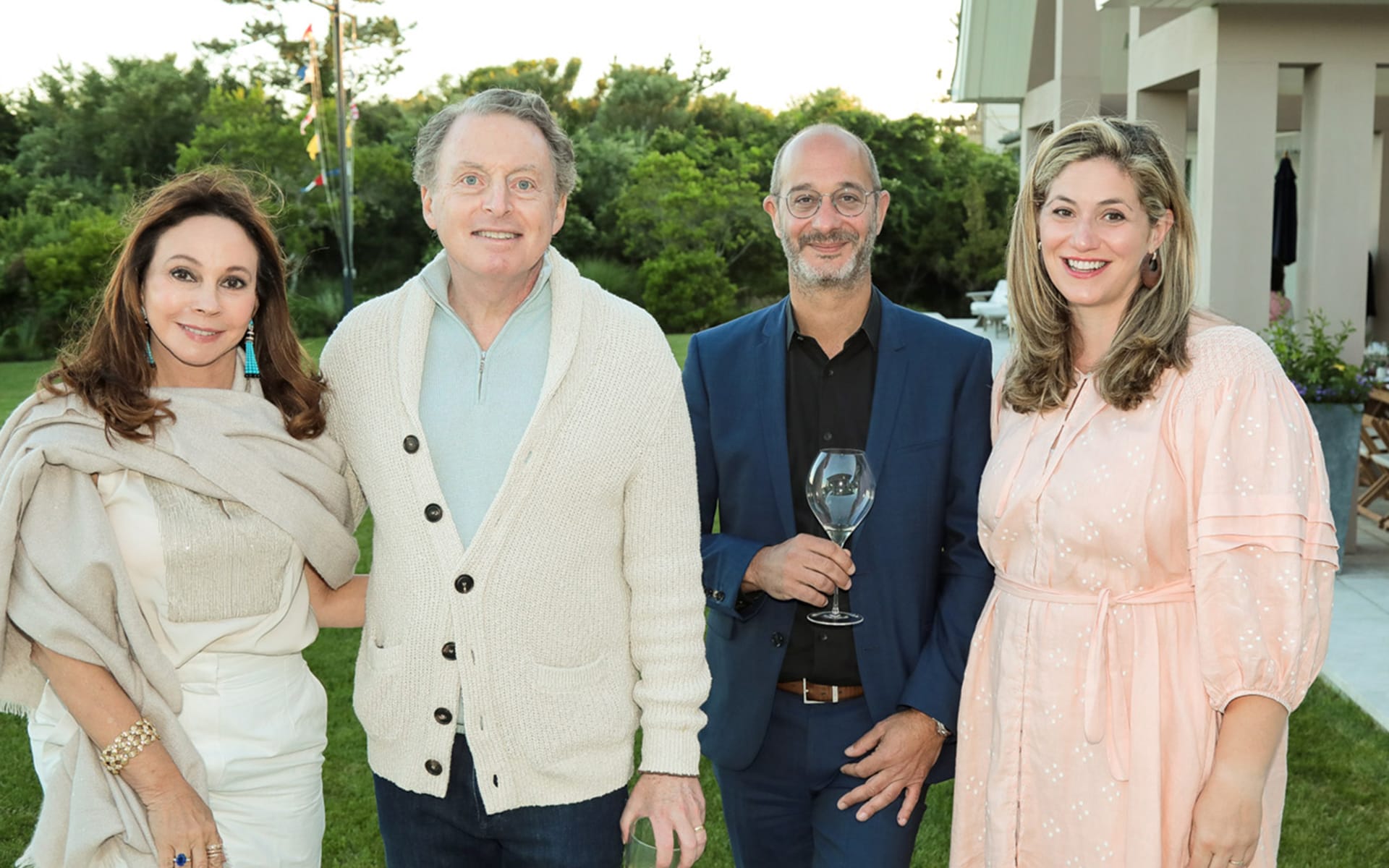 Four people standing outside with greenery and a house in the background, one holding a wine glass, all smiling.