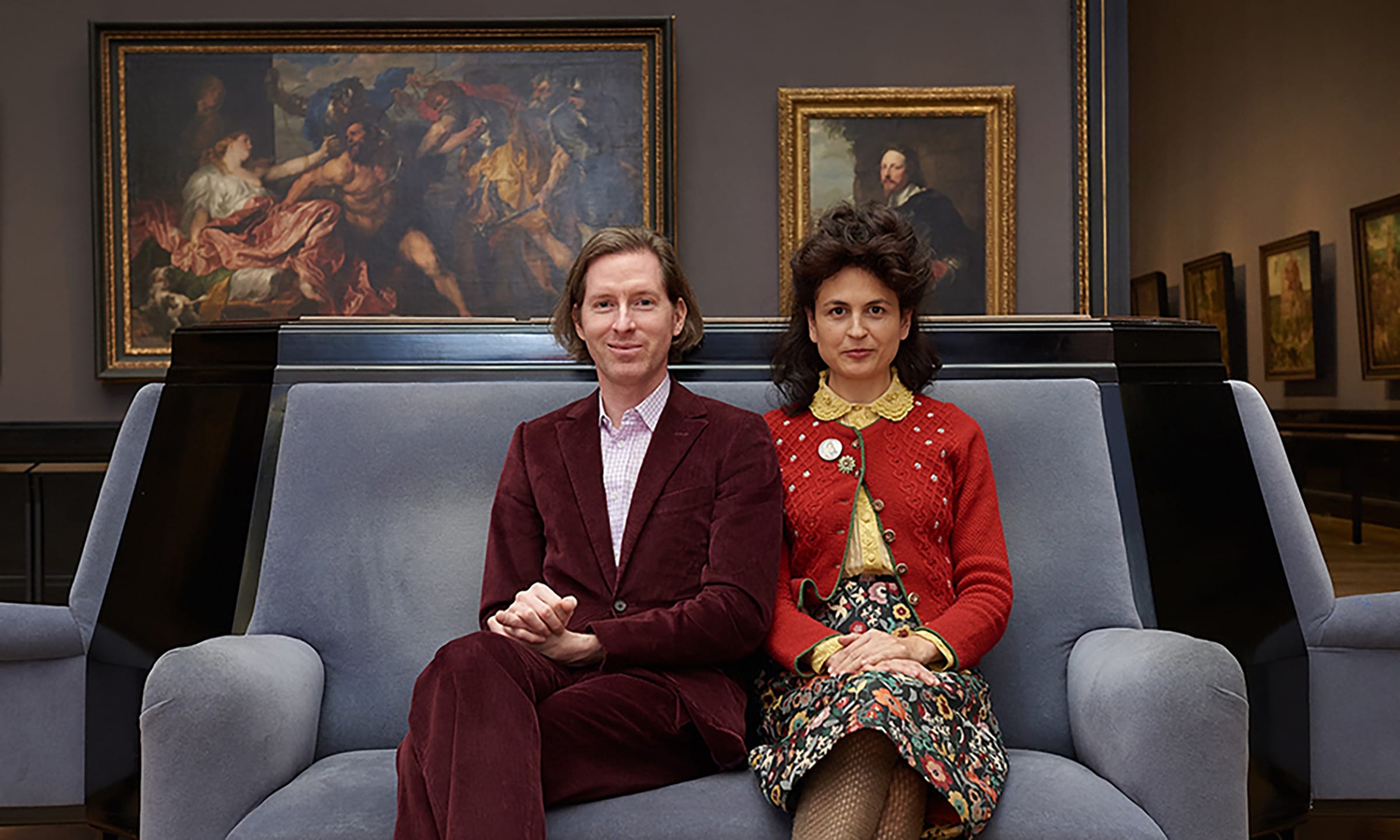 Two people sitting on a blue couch in an art museum gallery with framed paintings displayed on the wall behind them.
