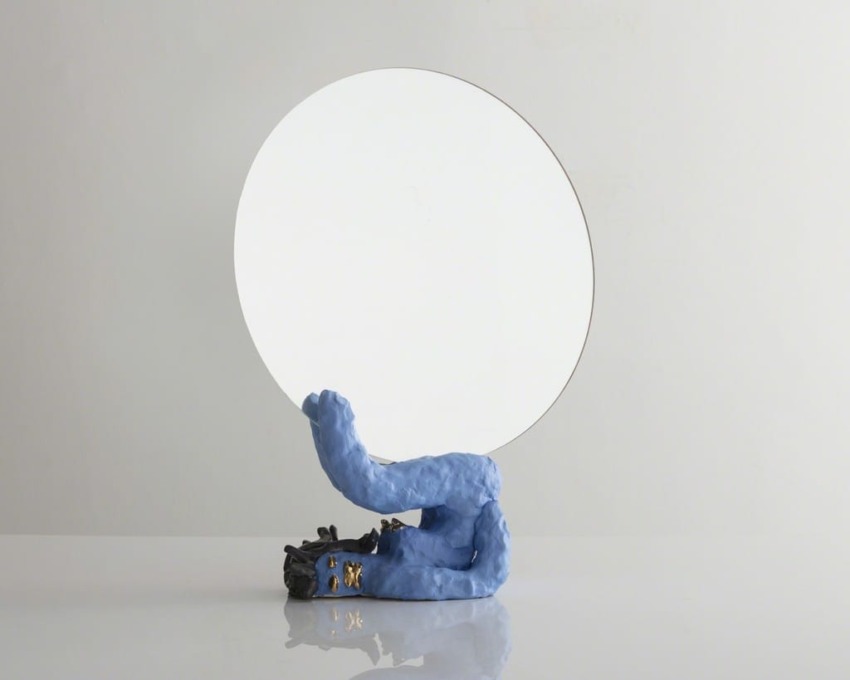 Round mirror with blue clay base on a reflective surface against a plain backdrop.