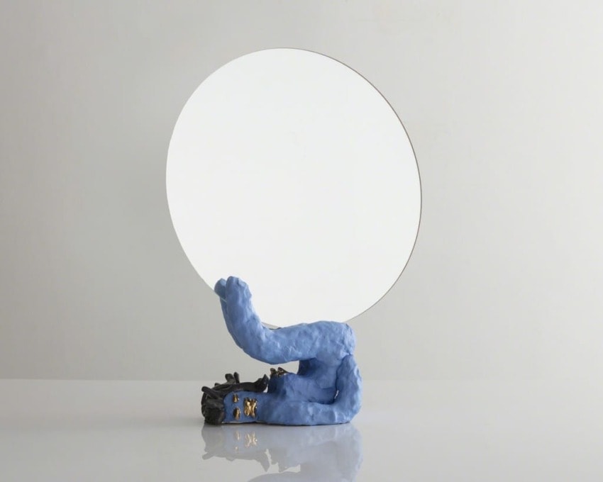 Round mirror with blue clay base on a reflective surface against a plain backdrop.