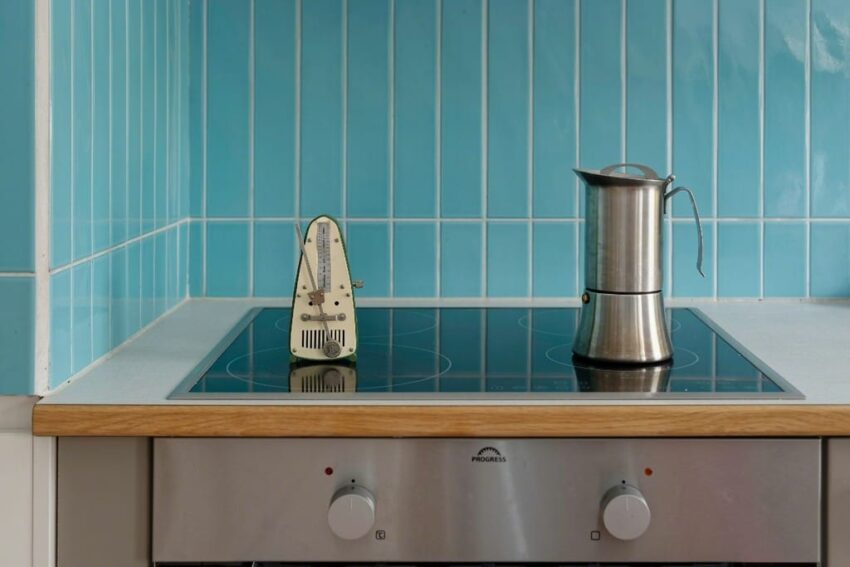 Stovetop with metronome and metal kettle against blue tiled wall in a kitchen setting.