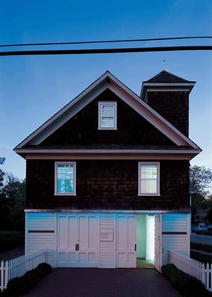 Evening view of a two-story house with illuminated door and windows, surrounded by a white picket fence.