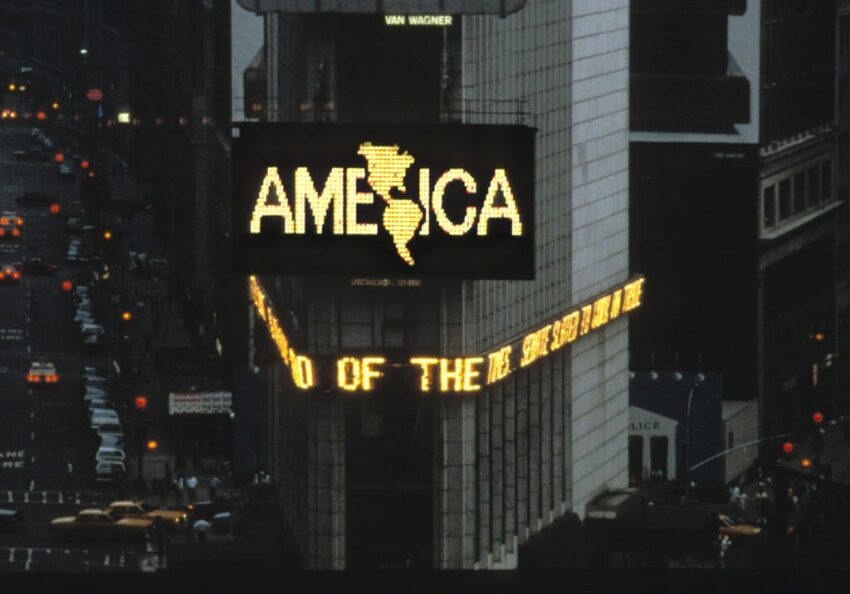 Times Square electronic billboard displaying "AMERICA" with city street view in the background.