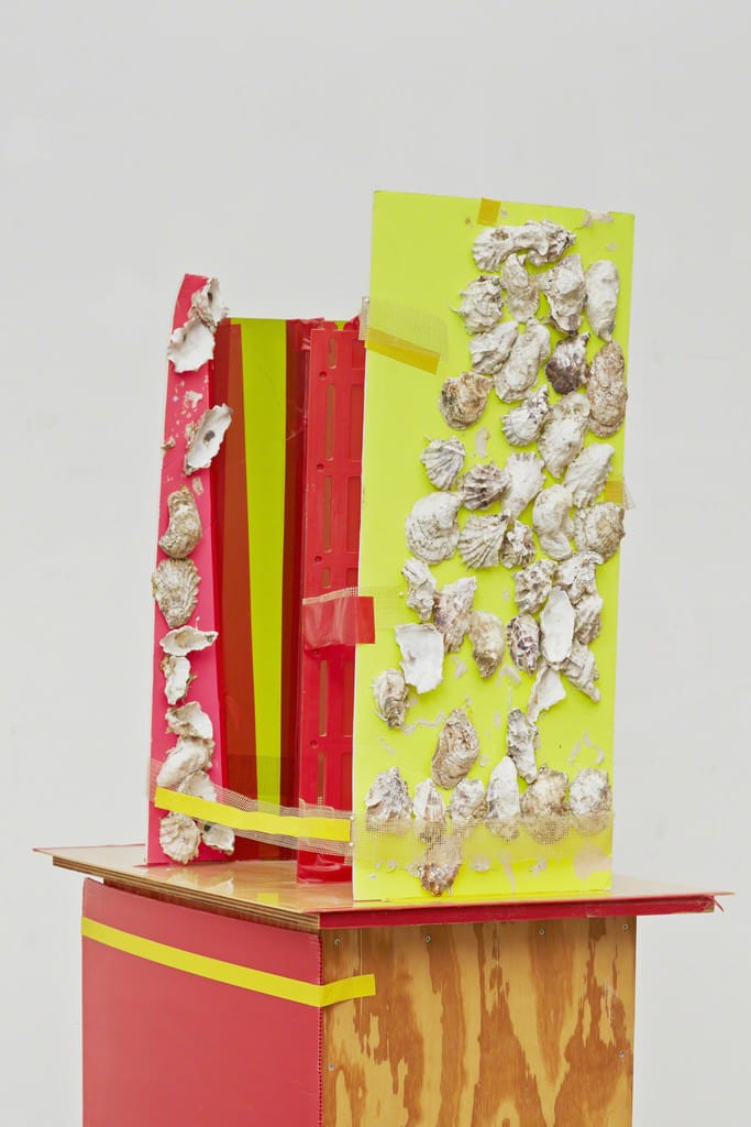 Abstract sculpture with red and yellow panels adorned with oyster shells, placed on a wooden base against a plain background.