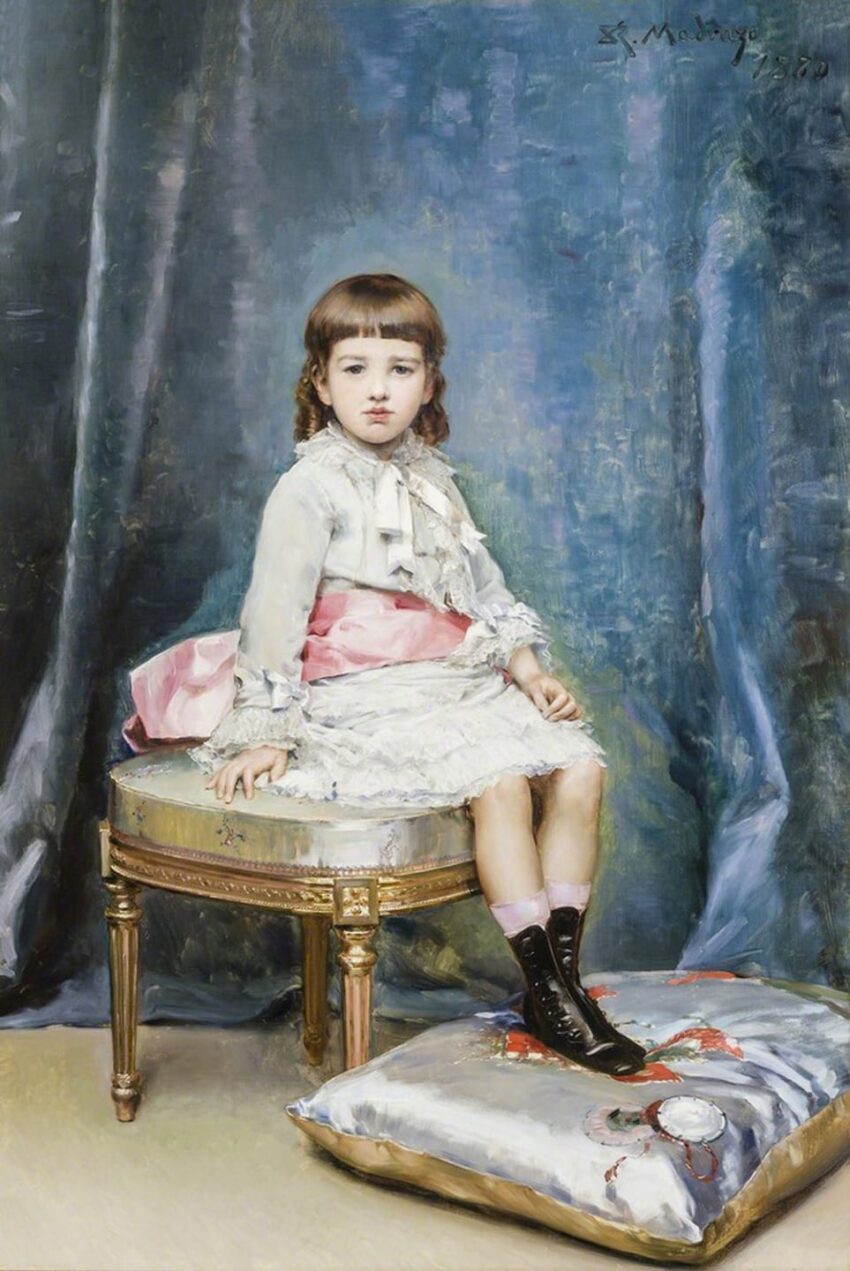 Young child in vintage attire sitting on a stool with blue drapery backdrop, wearing black boots and a dress with a pink sash.