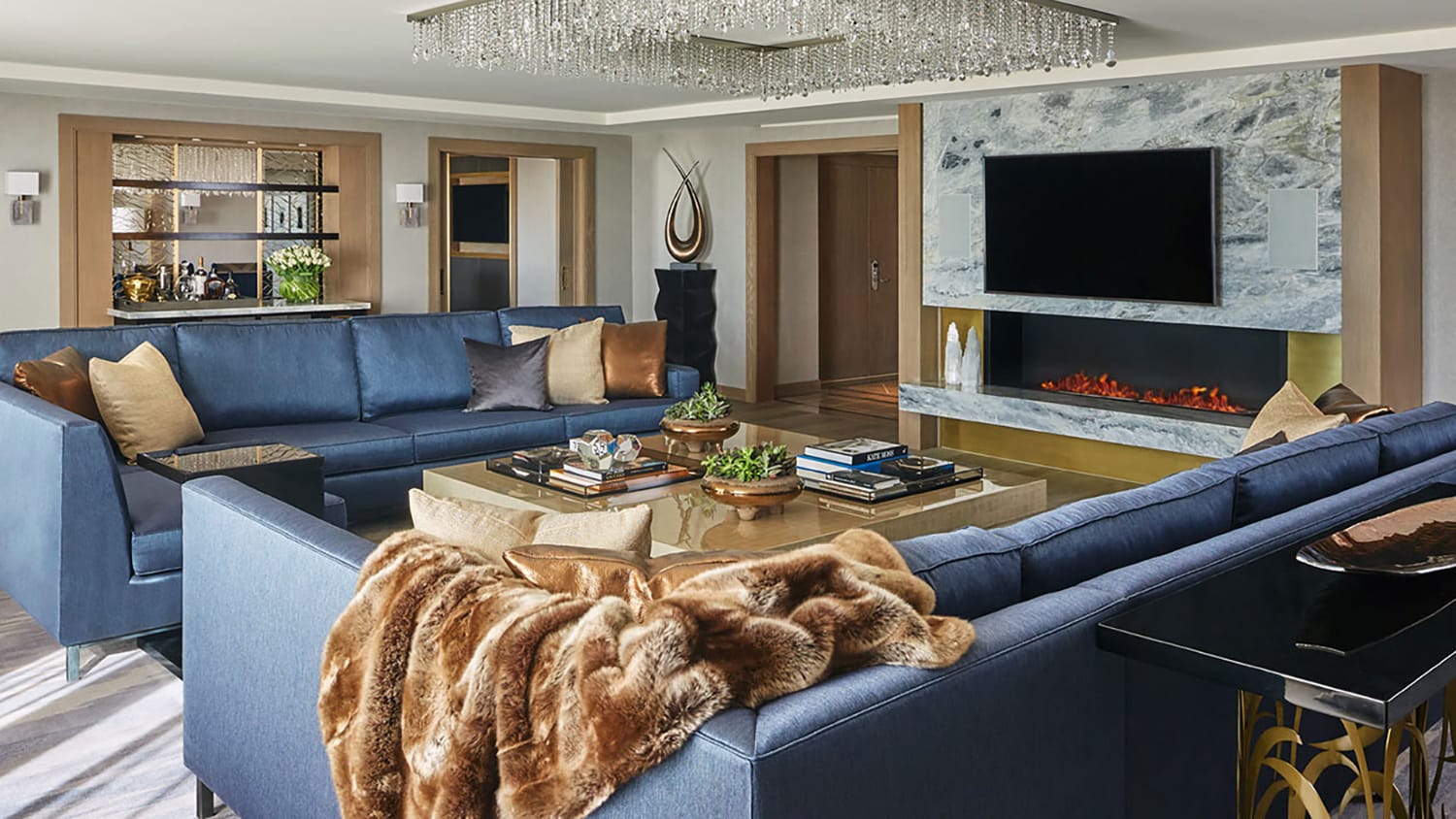 Luxurious living room with blue sectional sofa, elegant chandelier, large TV, modern fireplace, and decorative accents.