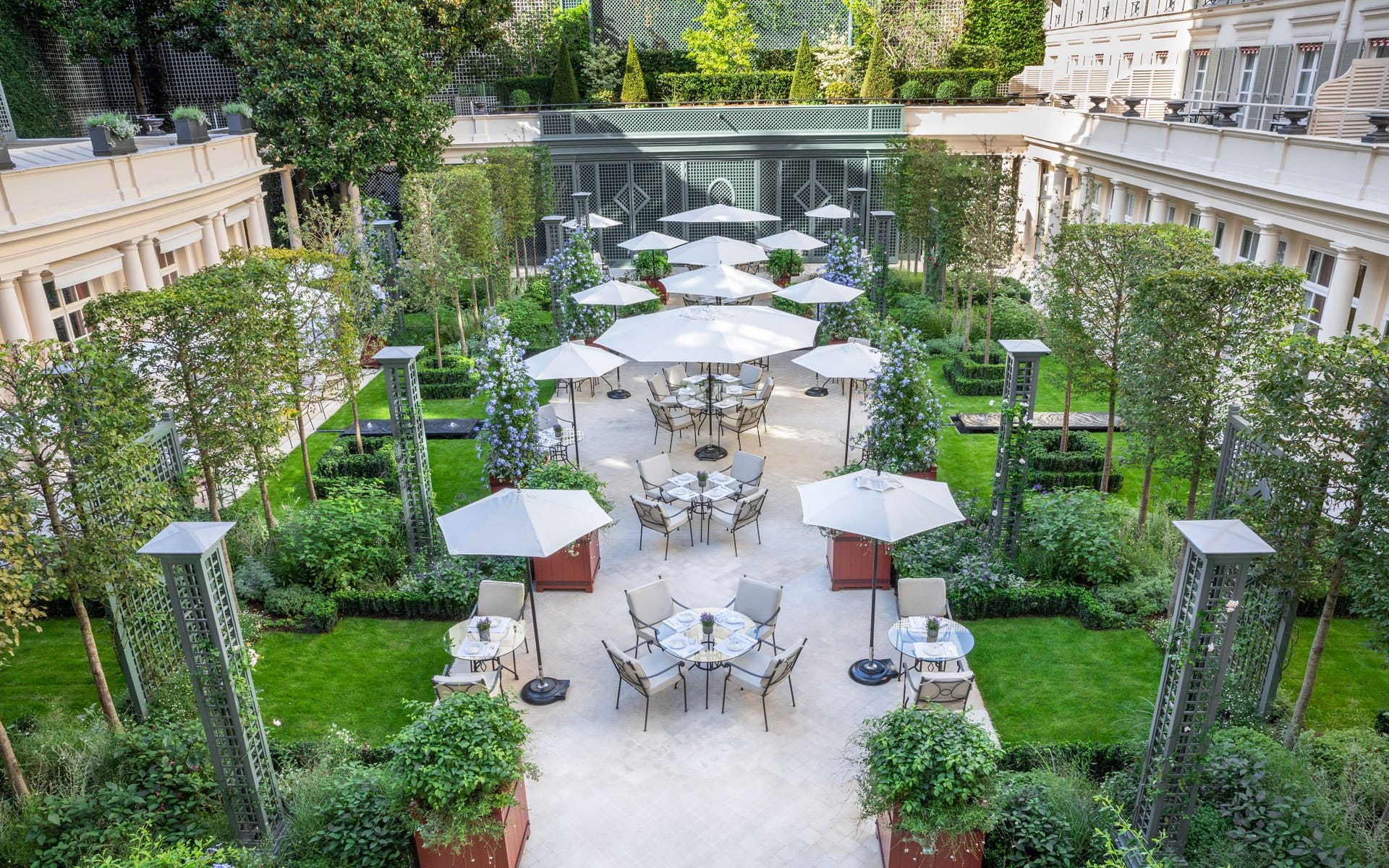Outdoor courtyard with tables, chairs, and umbrellas surrounded by lush greenery and tall buildings.