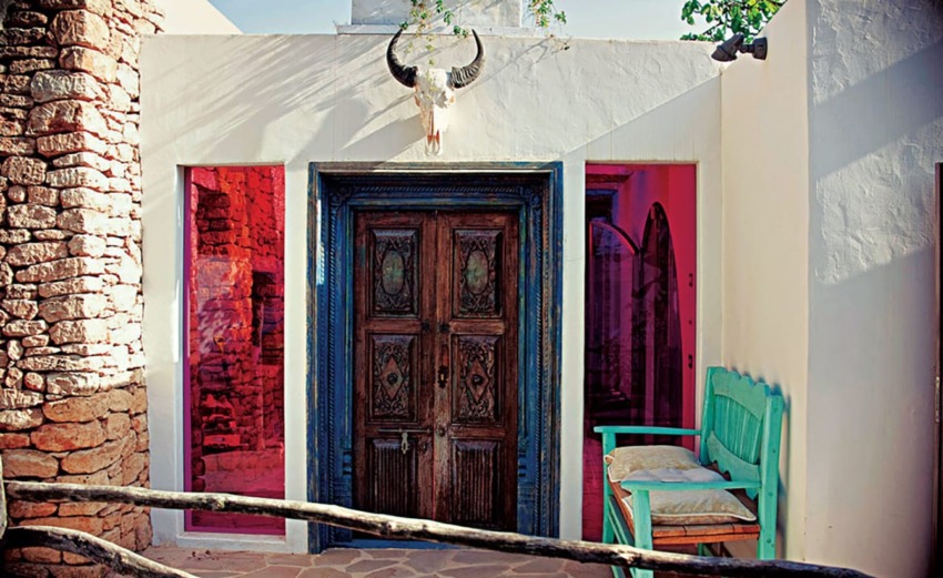 Vintage wooden door with skull decor, framed by bright pink windows and a turquoise bench in a rustic stone-walled setting.