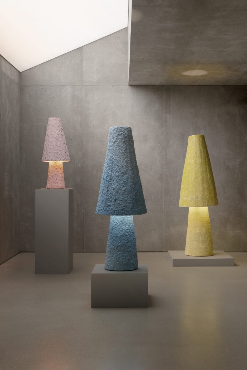 Three modern textured lamps on pedestals in a minimalist concrete room with soft lighting overhead.