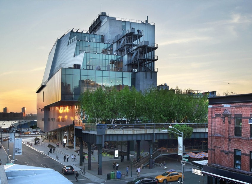 Modern art museum with glass facade, elevated platform, and trees at sunset, with people and taxis in the street below.
