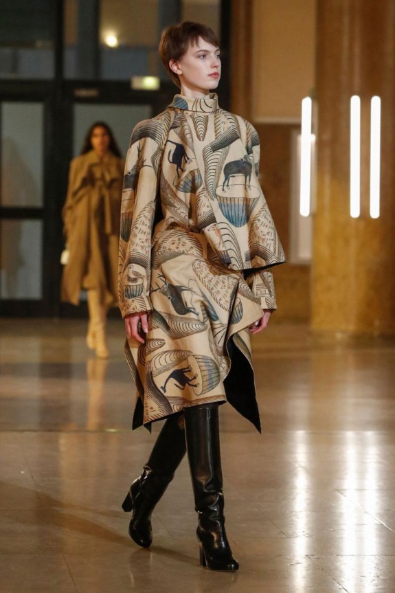Model wearing a patterned coat with animal designs, walking in a fashion show, with a second model in the background.