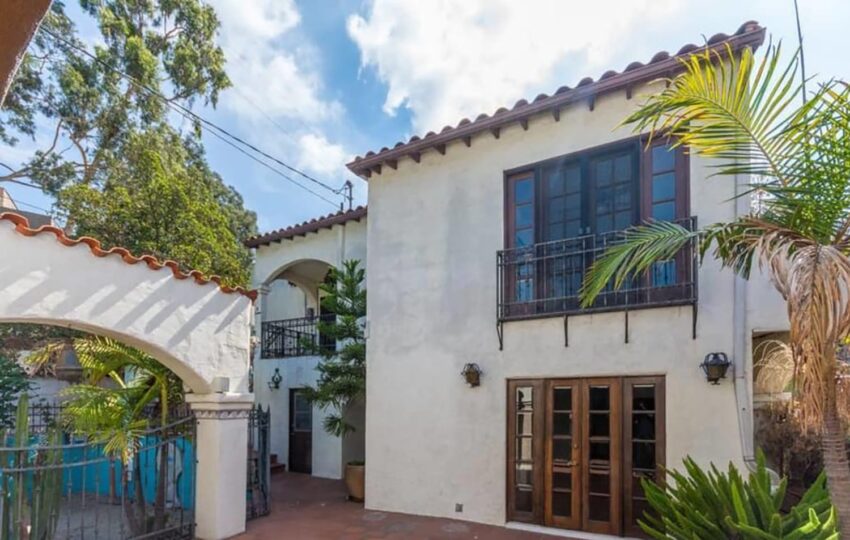 Mediterranean-style house with arched entrance, balcony, and palm trees in front, under a partly cloudy sky.