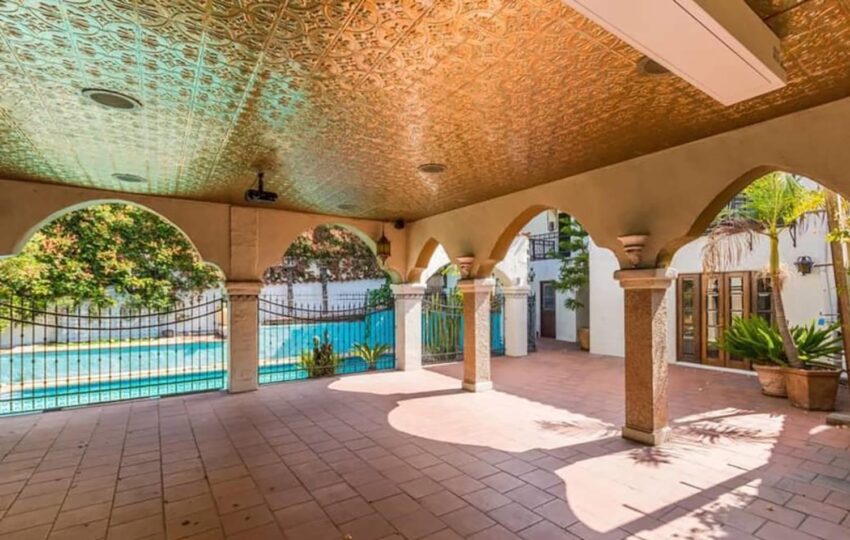 Covered outdoor patio with ornate ceiling, archways, and view of a pool surrounded by lush greenery and potted plants.