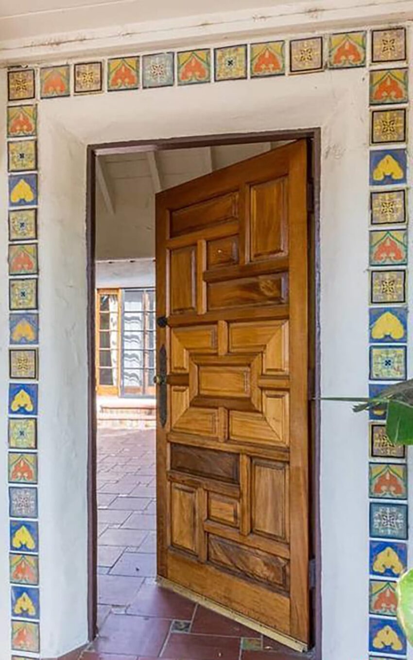 Wooden door with geometric patterns, surrounded by colorful decorative tiles, opens to a tiled interior space.