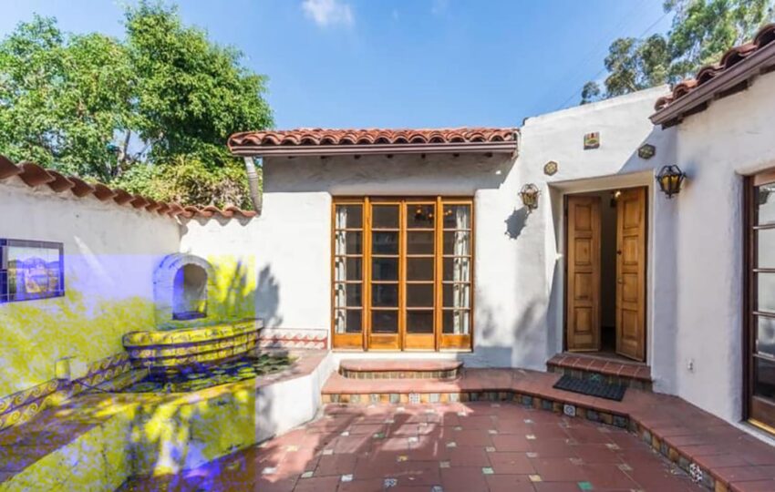 Spanish-style courtyard with tiled patio and wooden doors under a clay tile roof, surrounded by white stucco walls and greenery.