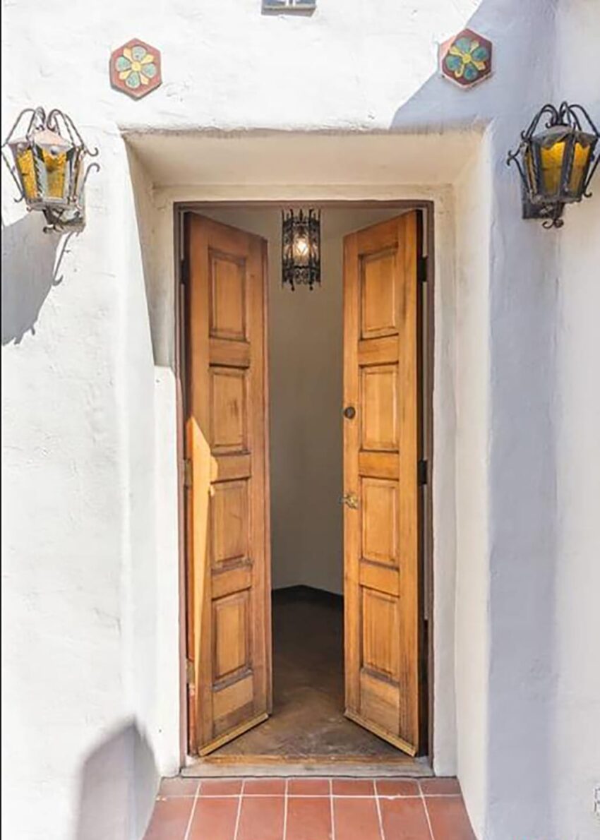Wooden double doors slightly open, with decorative lanterns on a white stucco wall.