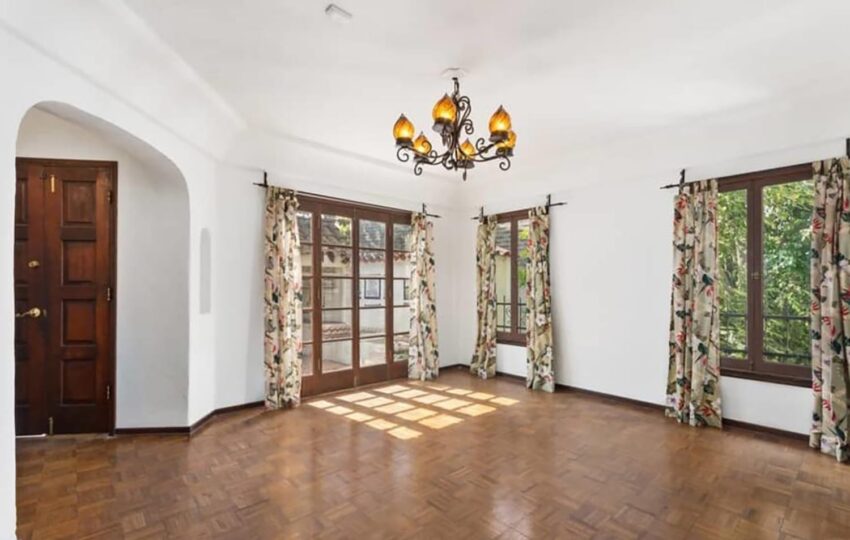 Spacious empty room with wooden flooring, floral curtains, large windows, and an ornate chandelier.