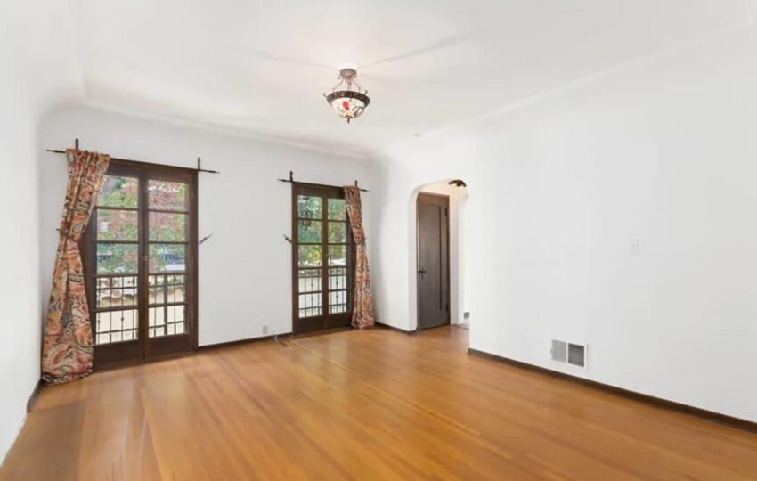Empty room with hardwood floor, white walls, large windows with floral curtains, and a decorative ceiling light fixture.