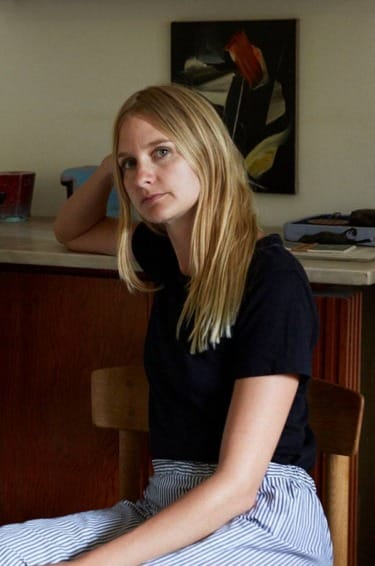 Person with long blonde hair sitting in a wooden chair, wearing a black shirt and striped pants, with a painting in the background.
