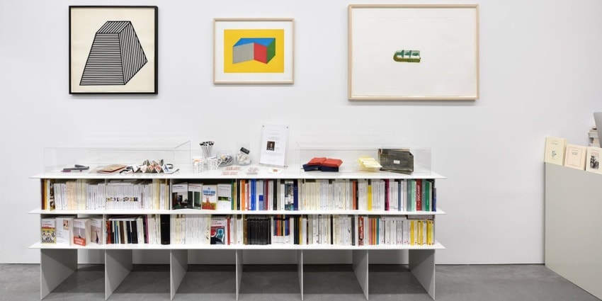 Art gallery display with framed geometric prints on the wall and a bookshelf filled with books and small items below.