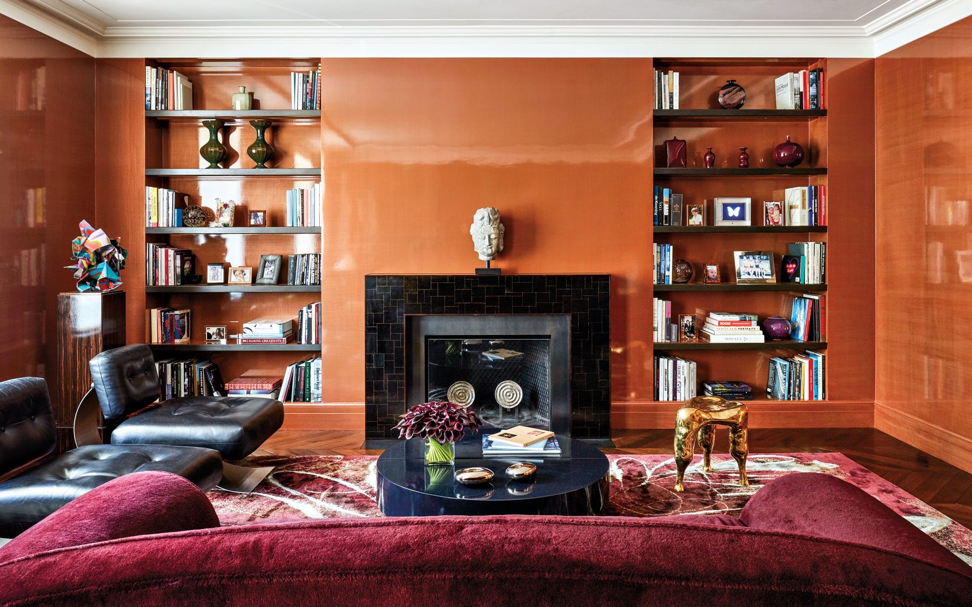 Stylish living room with orange walls, bookshelves, fireplace, black armchairs, red sofa, and decorative items.
