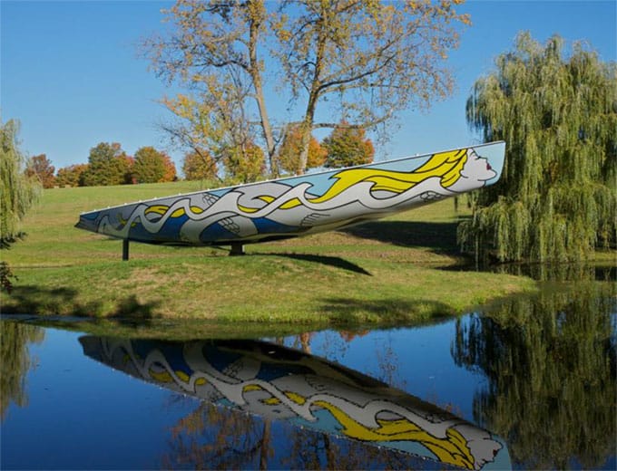 Colorful pop art sculpture of a canoe by a pond with trees and a clear blue sky in the background.