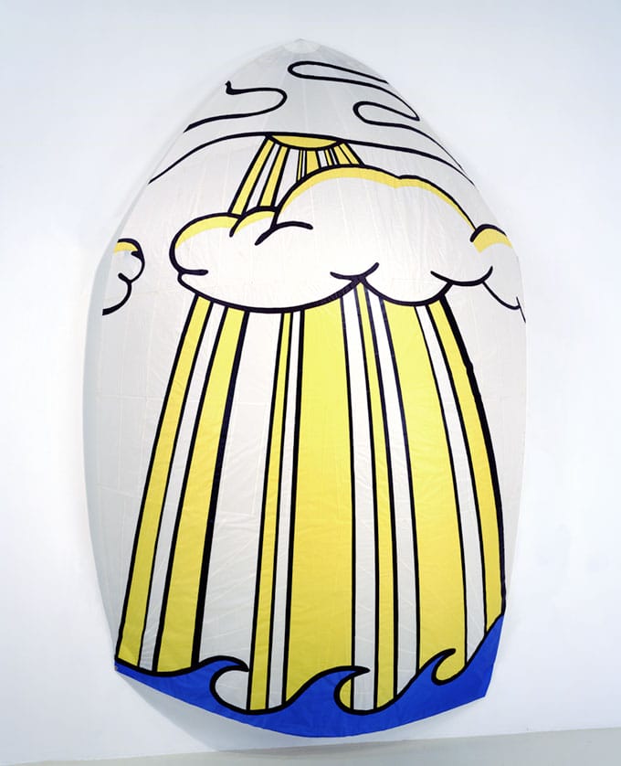 A surfboard painted with a pop art design featuring yellow sun rays, stylized clouds, and blue waves.