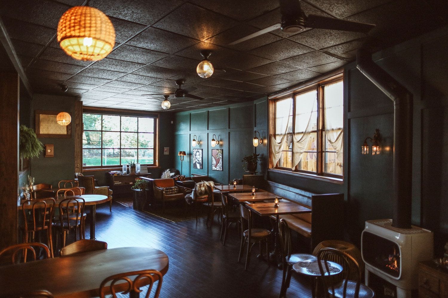 Cozy cafe interior with wooden tables, chairs, and a warm ambiance. Soft lighting from hanging lamps and large windows.