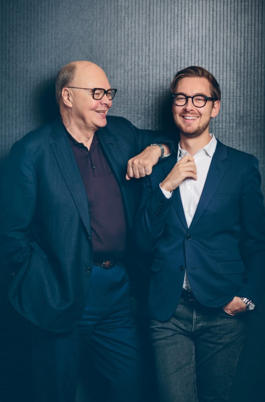 Two men smiling while standing against a dark textured backdrop, both wearing suits and glasses.