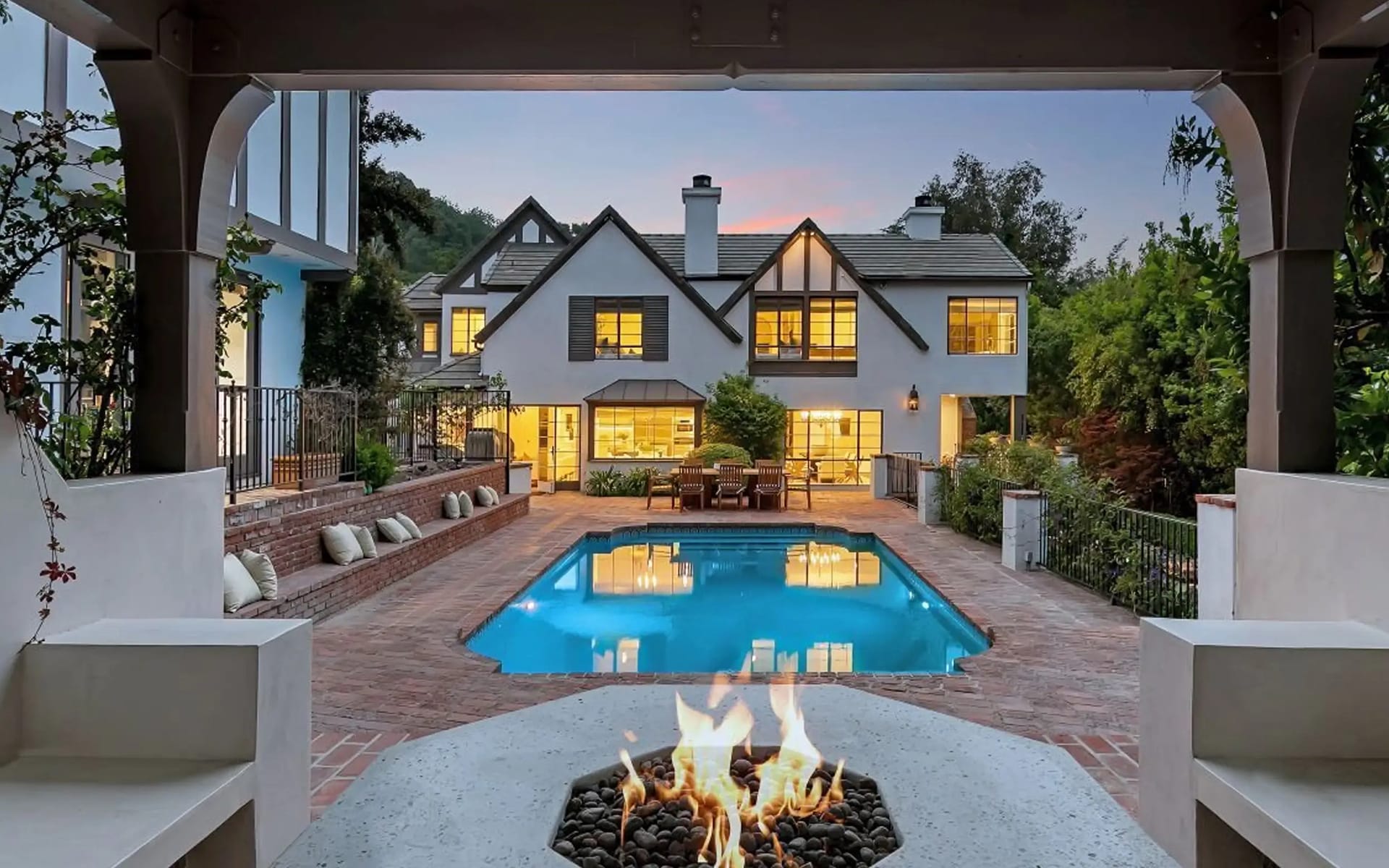 Luxury house exterior with lit windows and patio, featuring a pool and fire pit in the foreground at sunset.