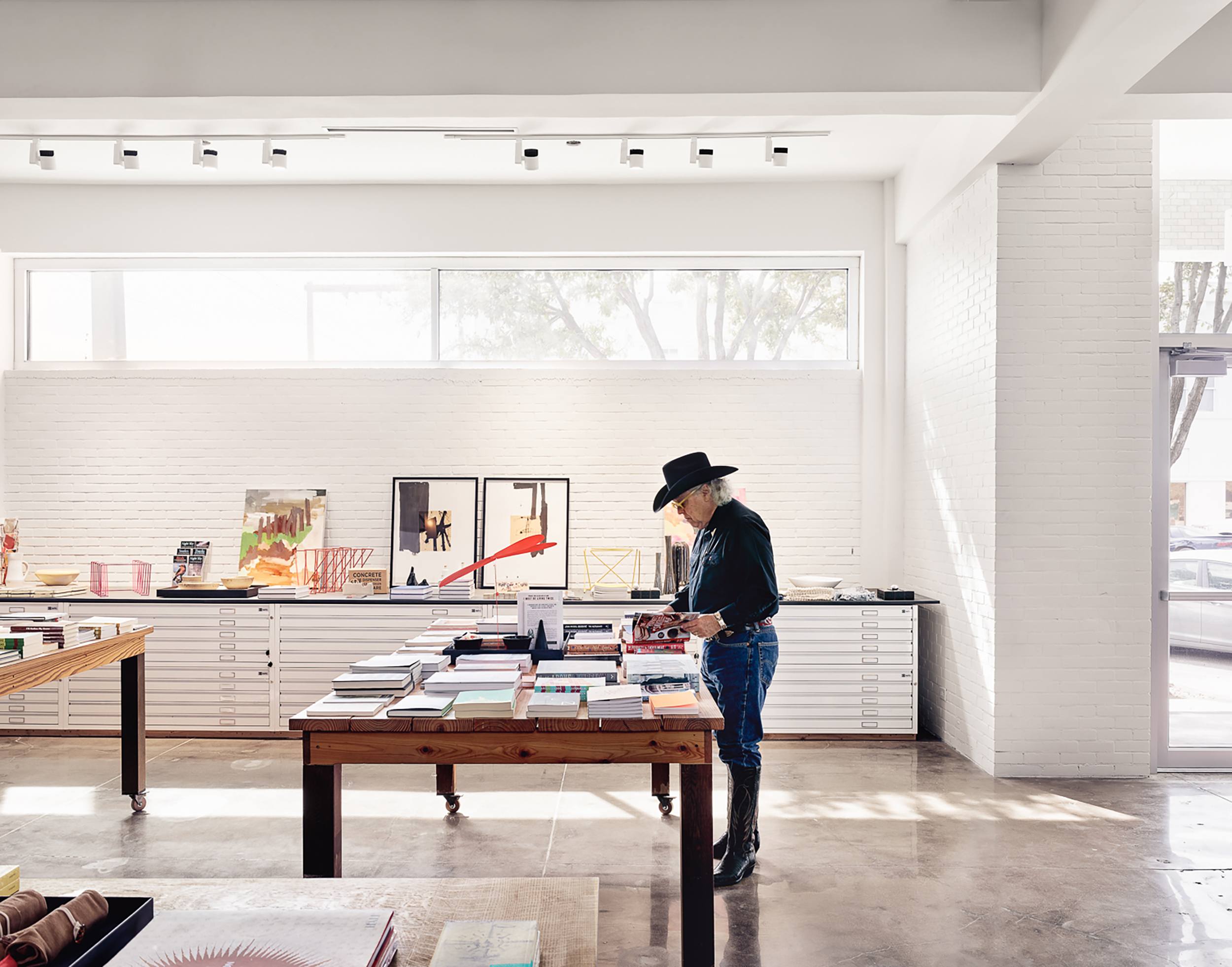 Person in a hat browsing books on tables in a modern, well-lit store with large windows and minimal decor.