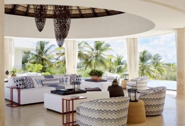 Luxurious open-air living room with white sofas, tropical decor, and view of palm trees and pool in sunny setting.