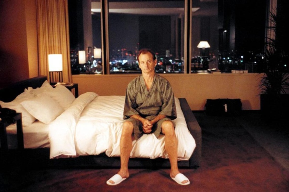 Man sitting on a bed in a dimly lit hotel room with a city view through a large window in the background.
