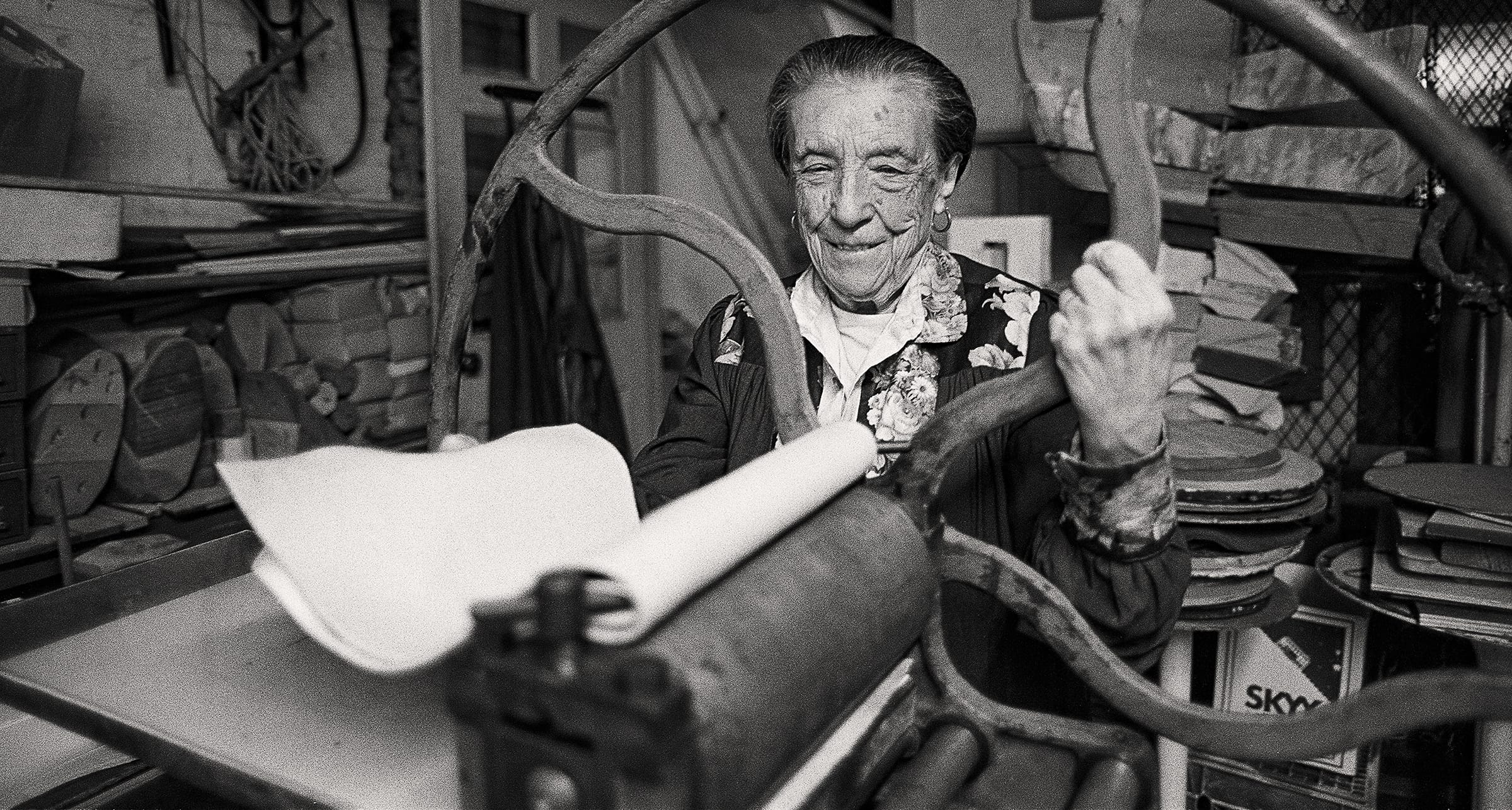 Elderly woman in a floral blouse smiling while operating a vintage printing press in a cluttered workshop.