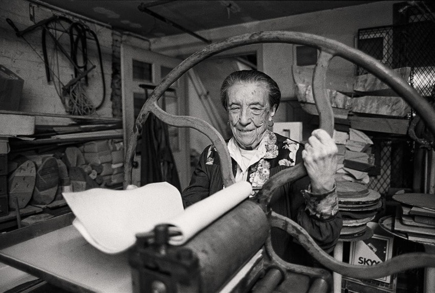 Elderly woman with a joyful expression operates an old printing press in a cluttered workshop filled with art supplies.