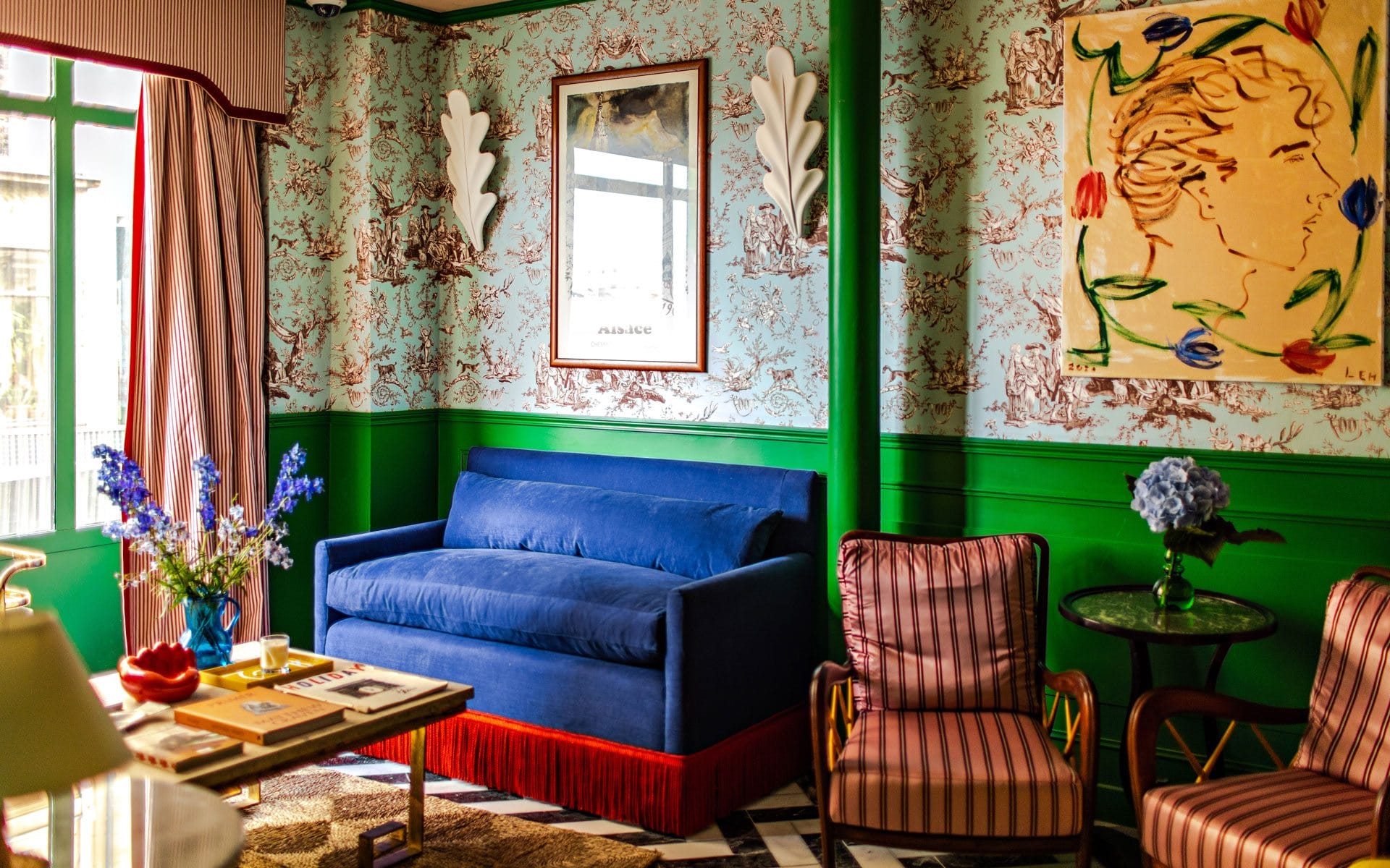 Colorful living room with blue sofa, floral wallpaper, artwork, striped chairs, and a coffee table with flowers and books.