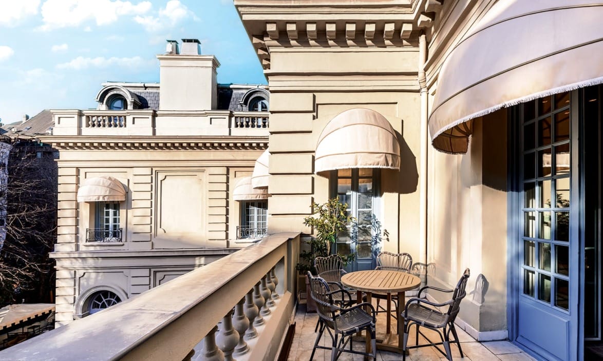 Elegant hotel balcony with table and chairs, overlooking classic architecture and blue sky.