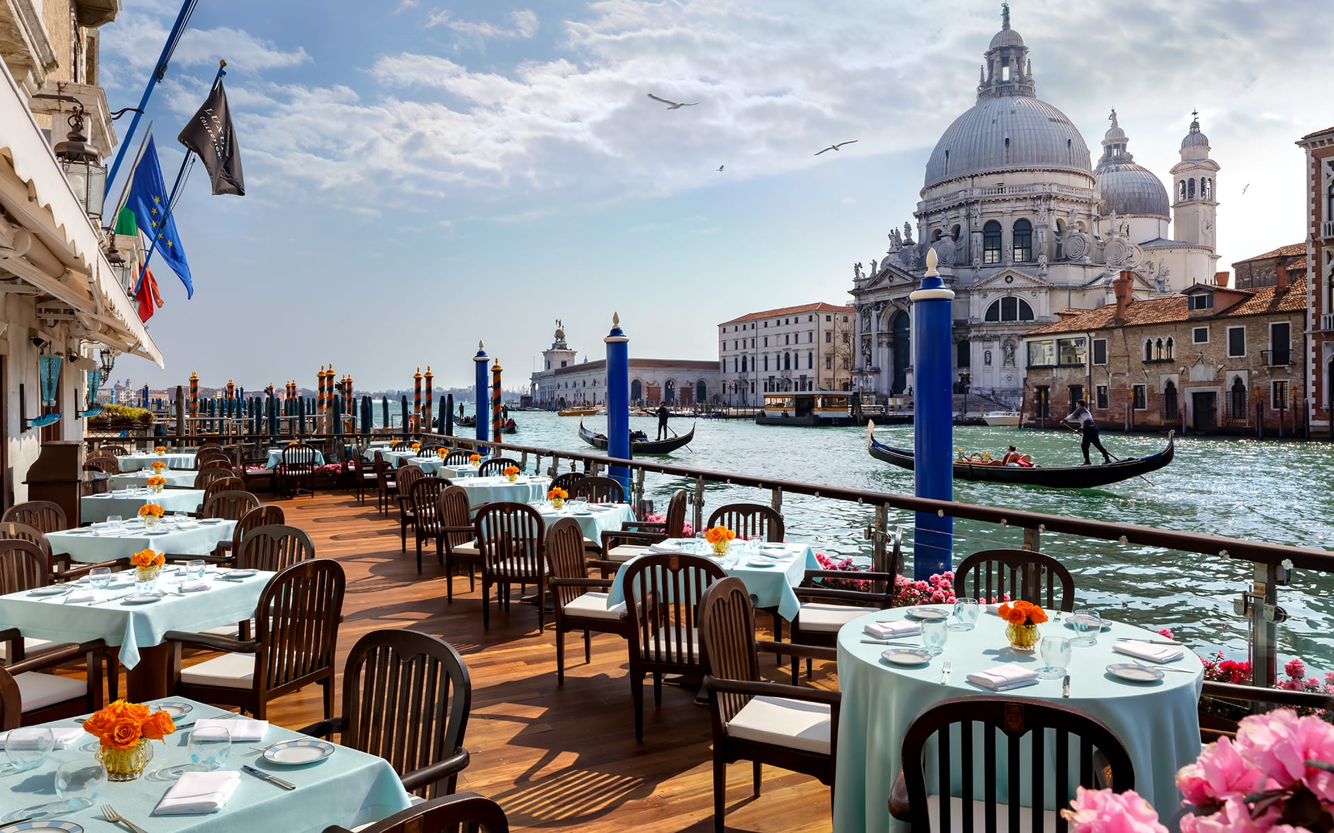 Outdoor restaurant terrace overlooking a canal with gondolas and historic architecture in Venice on a sunny day.