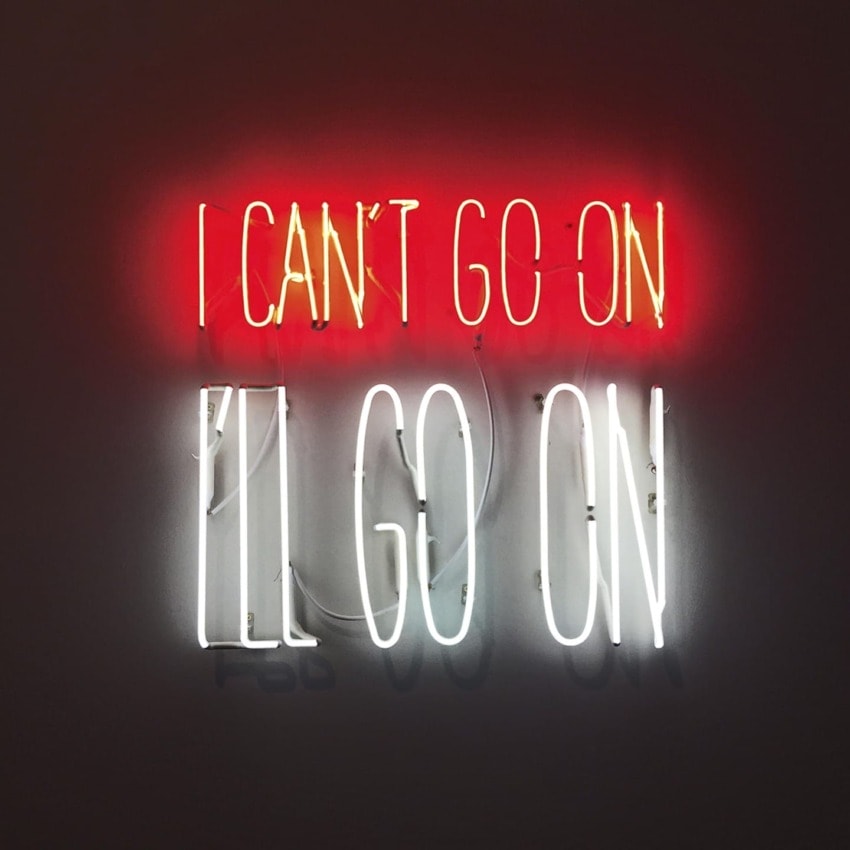 Neon sign with text "I can't go on I'll go on" in contrasting red and white lights on a dark background.