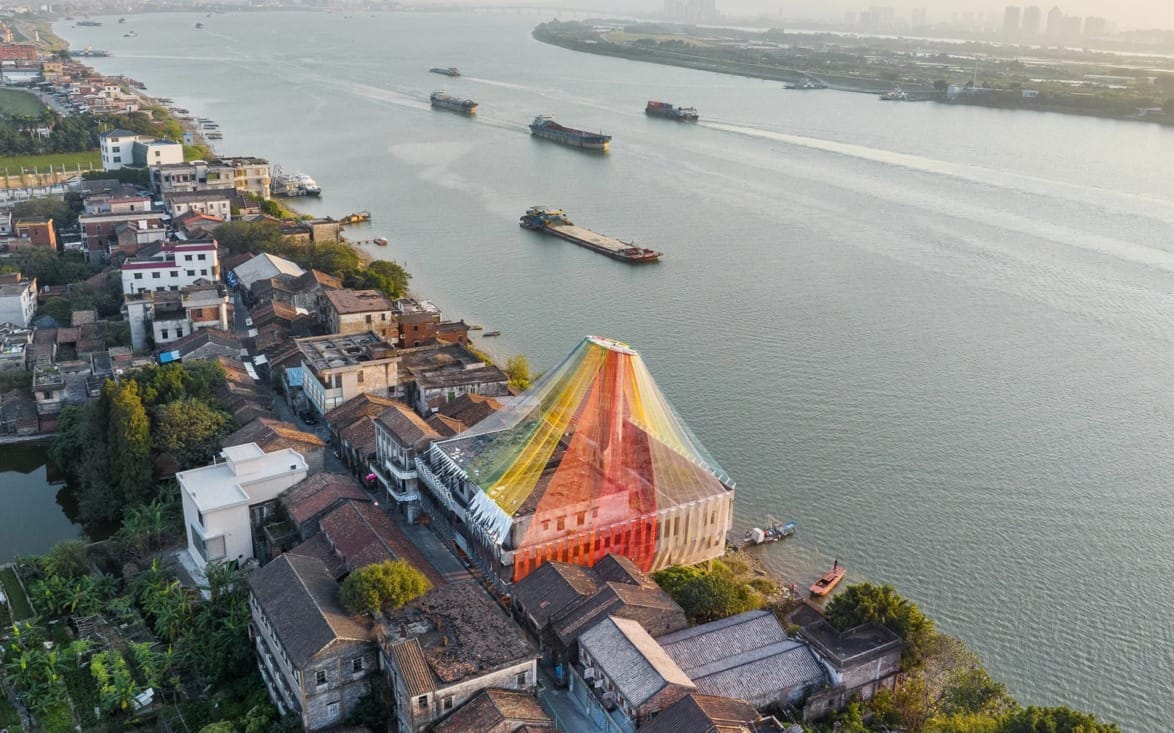 Colorful pyramid-roofed building by a river with boats, surrounded by an urban landscape and greenery at sunset.