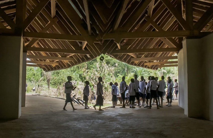 Group of children in uniforms gathering inside a wooden structure, with a view of trees outside in the background.