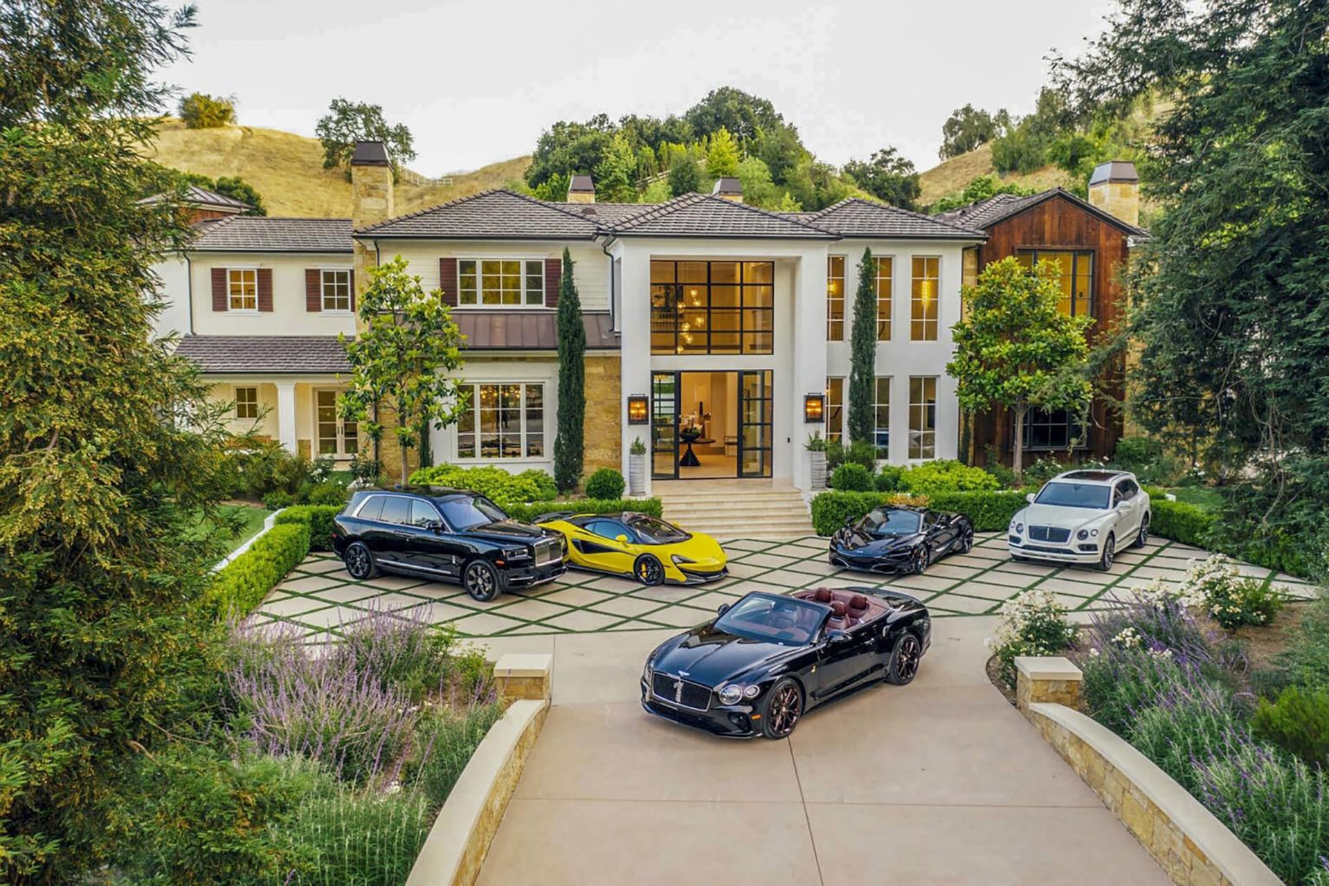 Luxury mansion with modern architecture, surrounded by exotic cars on a driveway, set amidst lush greenery.