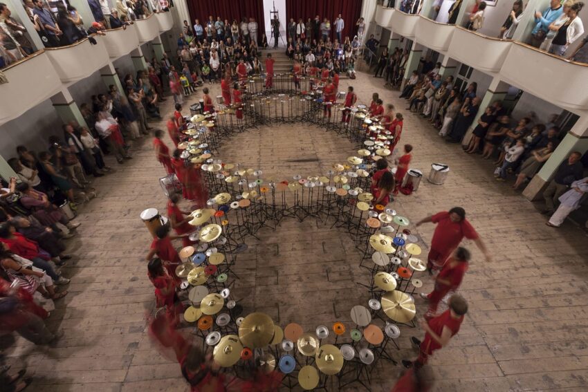 Aerial view of performers in red playing musical instruments in a circular formation before an audience in a large hall