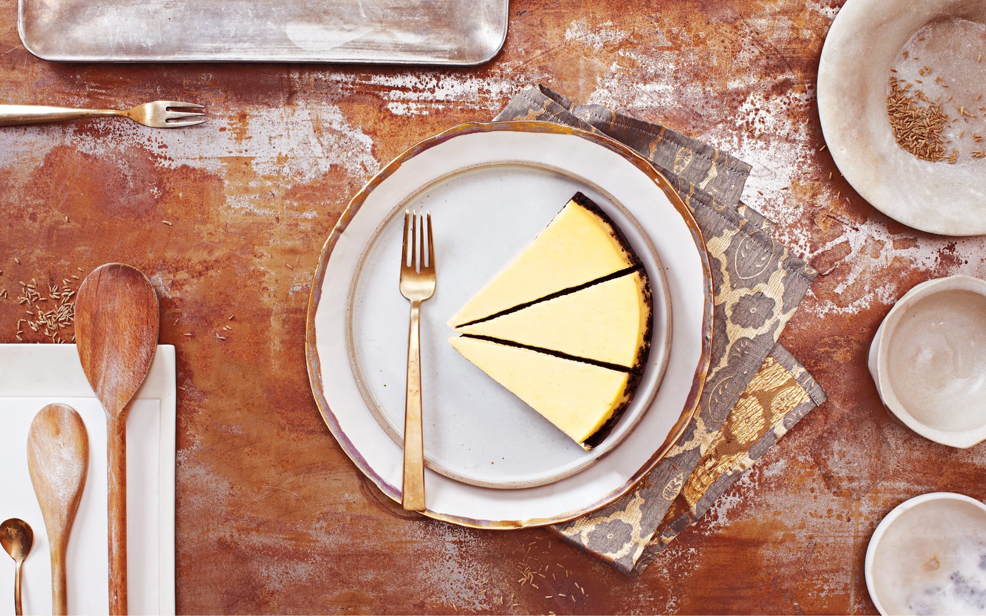Top view of a slice of cheesecake on a decorative plate with a gold fork, surrounded by various rustic kitchen utensils.